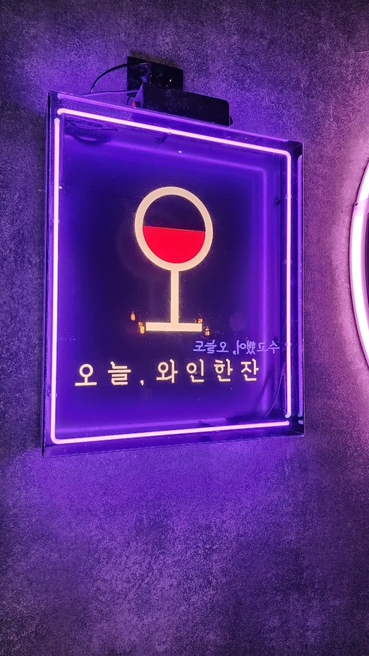 경상남도 진주시 오늘와인한잔 진주혁신점 보라색 네온사인 조명 아래 와인 잔 로고가 빛나는 벽면. 1인 좌석에서 즐기는 와인 혼술 상황.