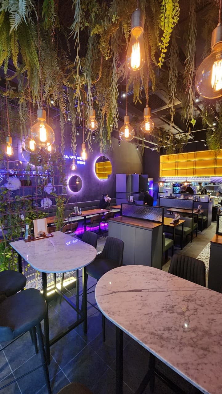 경상남도 진주시 오늘와인한잔 진주혁신점 와인바 - 천장 식물과 전구 조명 아래 대리석 테이블 및 혼술 가능한 보라색 네온의 다찌석과 테이블