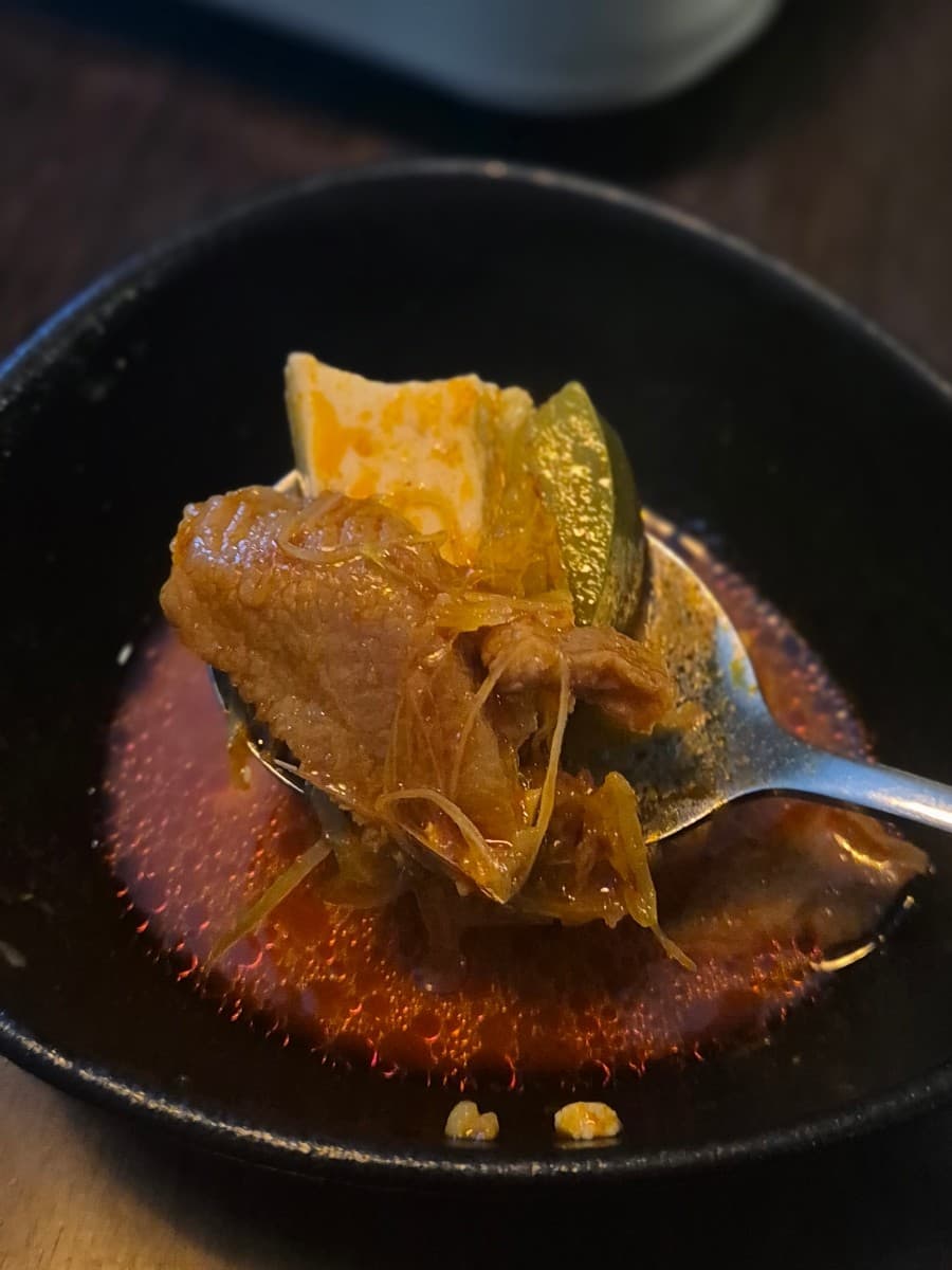 서울특별시 송파구 요리주점 조조모모 다찌석 1인 혼술 상황. 숟가락에 담긴 고기와 두부, 채소가 포함된 빨간 국물 요리와 검은색 그릇 위로 비치는 어두운 실내 조명.