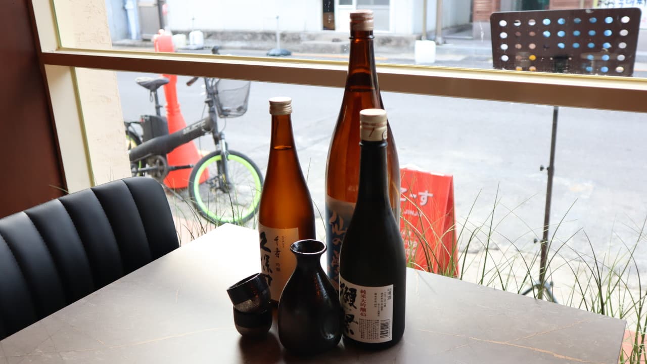 경상남도 진주시 오코스 진주칠암본점 진주 요리주점 창가 좌석. 테이블 위 사케 병들과 도자기 술병 및 잔이 놓인 1인 혼술 상황. 창밖 거리 풍경이 보이는 실내 좌석 구성.