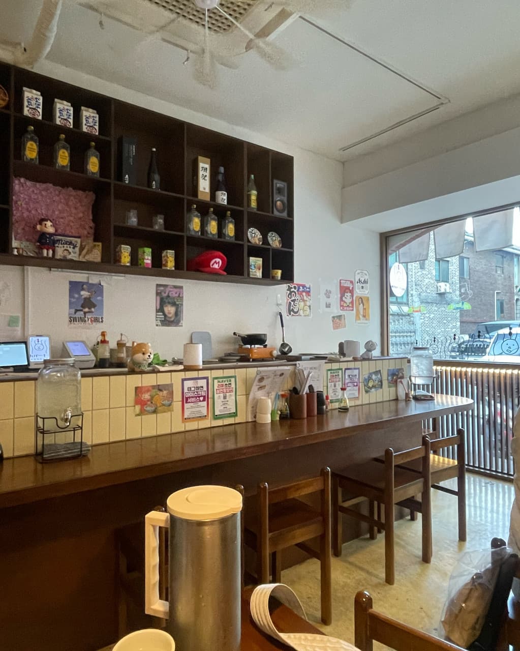 서울특별시 송파구 오뎅바모츠뎅 잠실 이자카야 내부 나무 소재의 1인 다찌석과 선반 위 진열된 사케병. 벽면 포스터와 밝은 창가 옆 혼술 공간의 배치 상황.