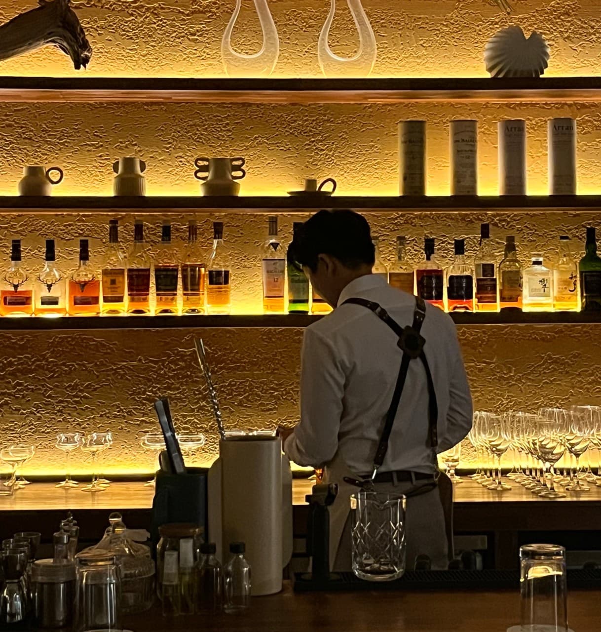 경상남도 진주시 혼술 바 슈럽 진주 - 노란 간접 조명이 비치는 질감 있는 벽면과 진열된 위스키 병, 1인 다찌석 카운터 앞에 선 바텐더