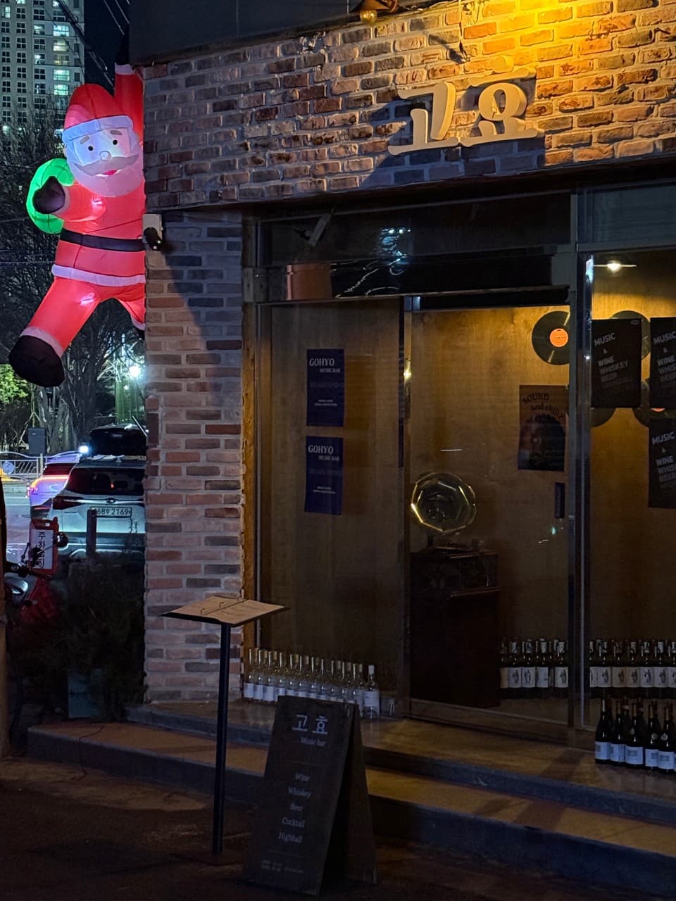 경상남도 진주시 혼술 와인 바 고효 진주. 벽돌 외관에 산타 장식이 설치된 입구와 노란 조명 아래 진열된 와인병, LP판이 보이는 실내 다찌석 분위기.