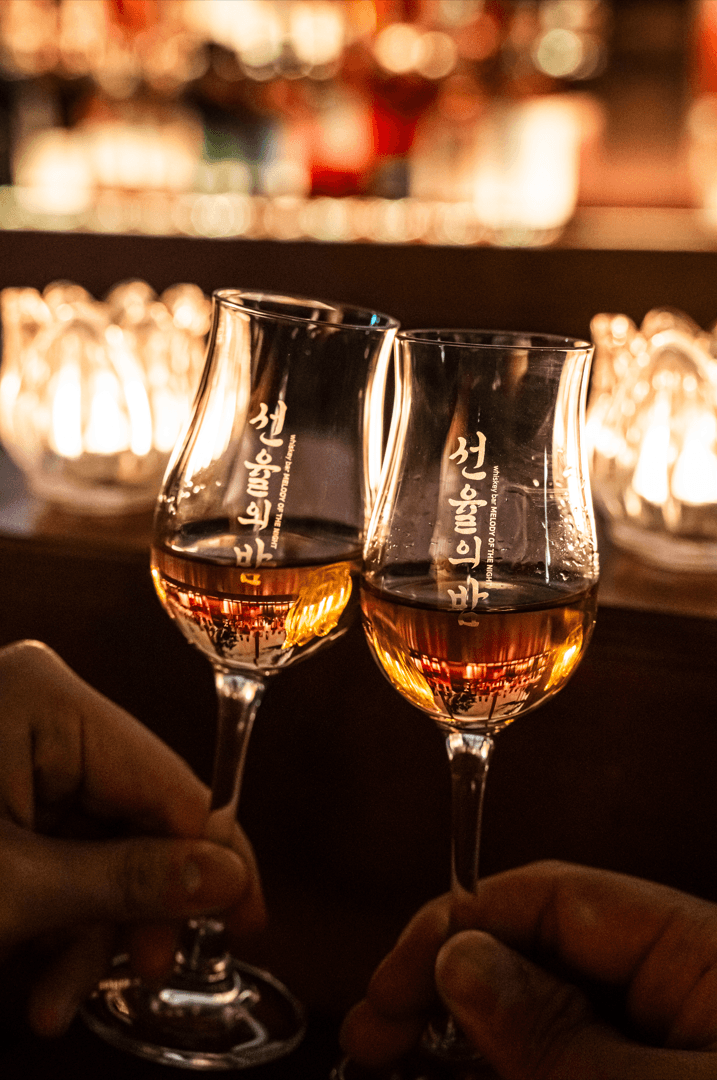 서울특별시 송파구 선율의밤 잠실방이점 바(Bar) 다찌석 조명 아래 위스키 잔을 든 상황. 투명한 글라스 속 호박색 액체와 빛이 번지는 배경이 혼술 분위기를 나타냄.