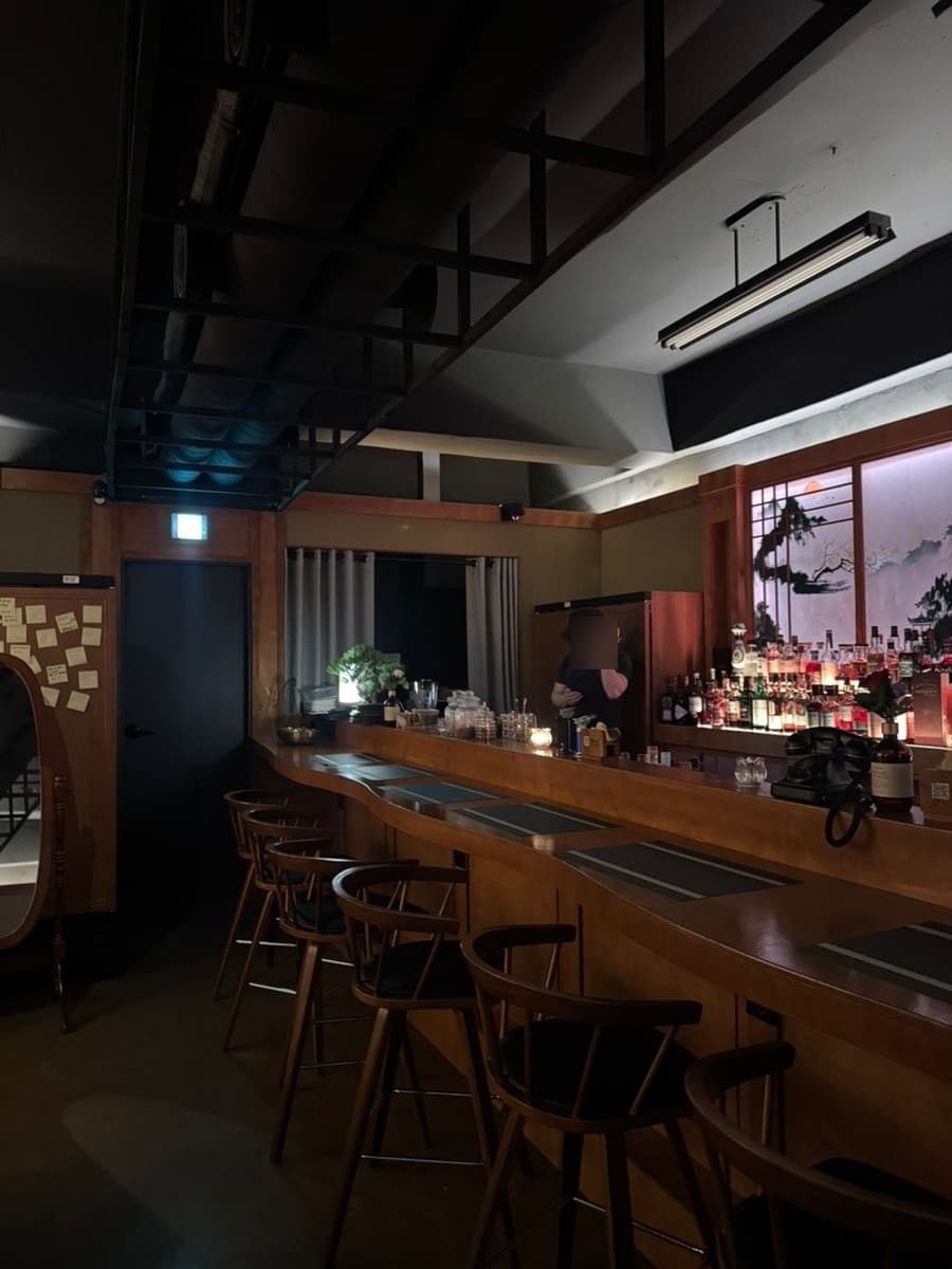 서울특별시 송파구 선율의밤 잠실방이점 바(Bar). 낮은 조명 아래 매끄러운 목재 다찌석과 1인석 의자가 배치된 혼술 공간. 배경의 수많은 술병들과 동양적 화풍의 조명이 비치는 내부.