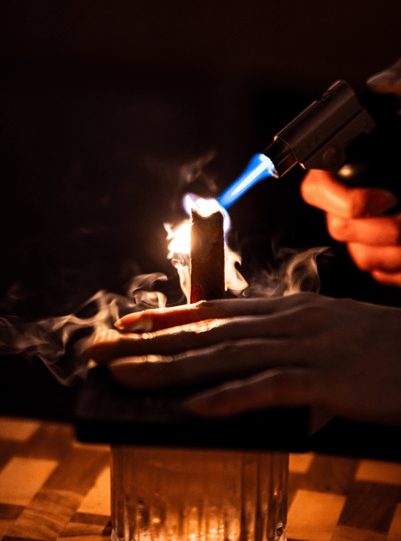 서울특별시 송파구 선율의밤 잠실방이점 바 다찌석 토치로 시나몬을 태우는 1인 혼술 칵테일. 어두운 조명 아래 피어오르는 연기와 술잔이 놓인 나무 테이블 위 칵테일 제조 과정.