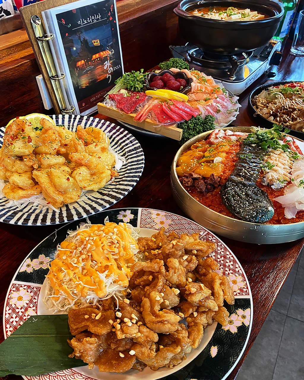 경상남도 진주시 미노리인사 진주 이자카야 나무 다찌석에 차려진 사시미와 튀김. 조명 아래 1인 혼술을 위한 해산물과 나베 요리의 질감이 돋보임.