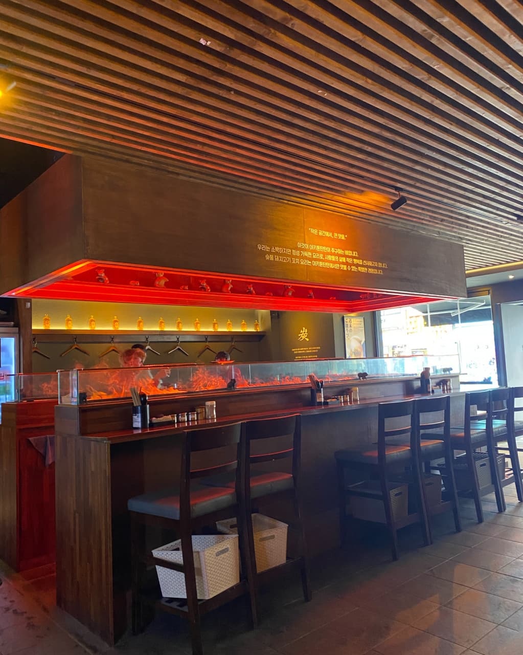 서울특별시 송파구 야키톤탄탄 잠실 이자카야의 붉은색 간접 조명과 1인 다찌석. 혼술 좌석으로 구성된 긴 카운터 테이블과 나무 소재의 천장 인테리어.