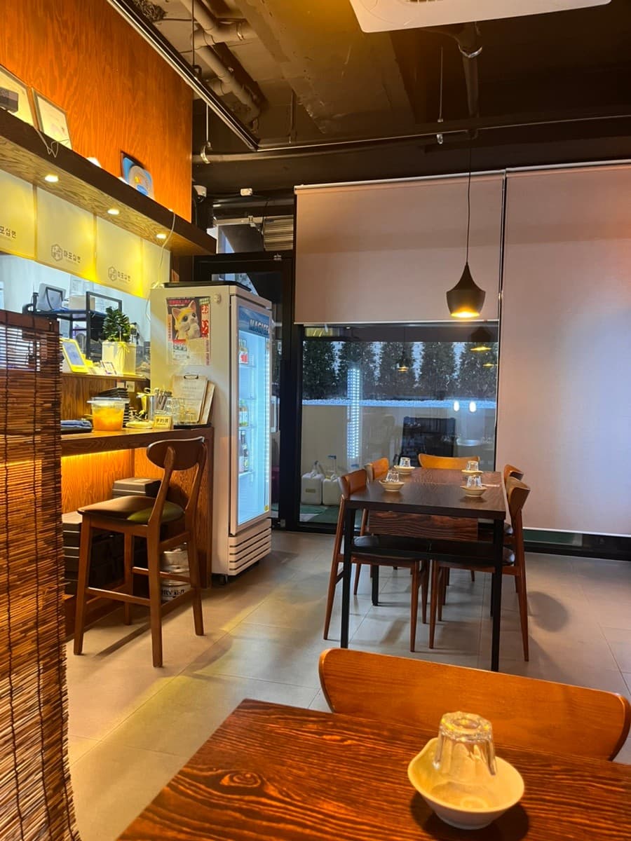 대전광역시 서구 이자카야 마포십번 대전서구의 노란 조명이 비치는 1인 다찌석과 목재 테이블. 혼술 하기 적당한 카운터 좌석과 창밖 풍경이 보이는 깔끔한 내부 구성을 보여줌.