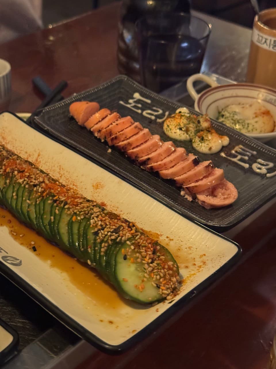 대전광역시 유성구 요리주점 도리토리 대전봉명점 다찌석에 차려진 명란구이와 오이. 낮은 조명 아래 1인 혼술 좌석과 안주의 질감을 강조함.