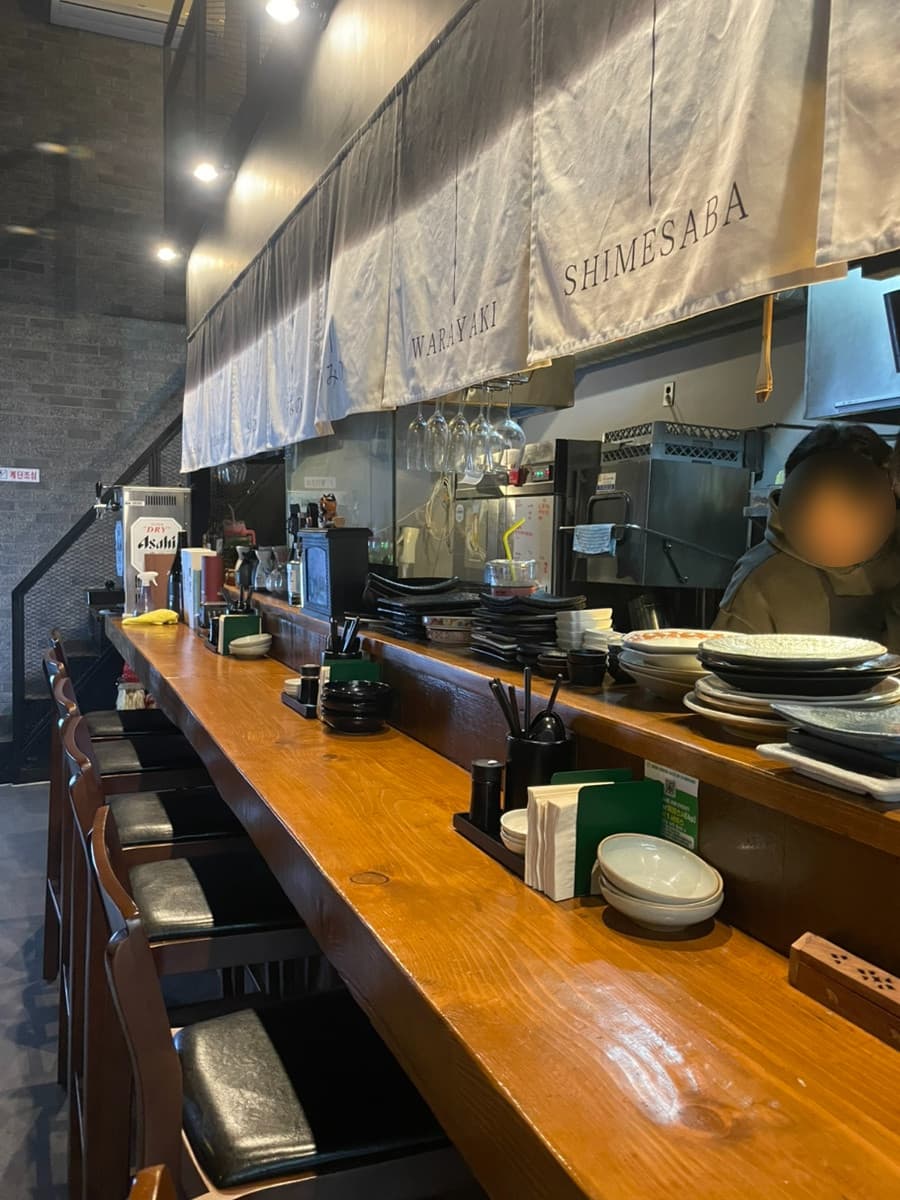 대구광역시 중구 이자카야 삼덕동 우미노. 조명이 비추는 나무 다찌석과 1인용 의자가 정렬된 내부. 시메사바 천막이 걸린 카운터에서 혼술을 즐기는 상황.