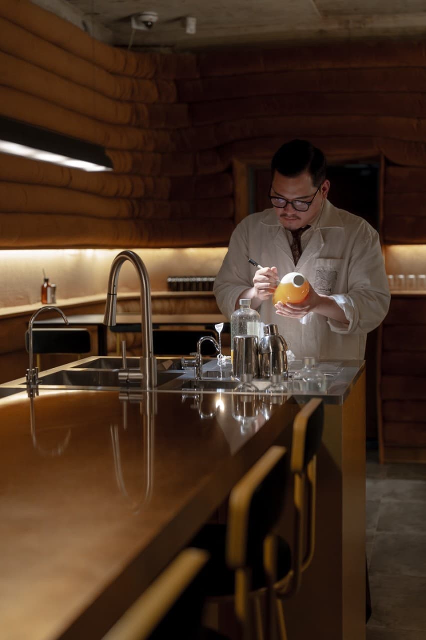 부산광역시 수영구 코듀로이 벨라즈 광안리 바(Bar)의 낮은 조도 아래 1인 다찌석과 음료를 준비하는 바텐더. 혼술에 최적화된 카운터석과 입체적인 벽면 질감이 돋보이는 내부 전경.