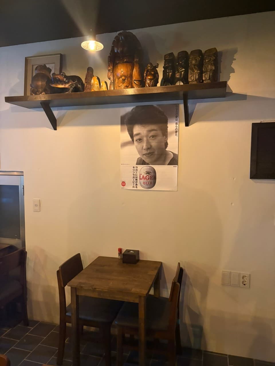 부산광역시 동구 이자카야 아지센 초량. 노란 조명 아래 2인 목재 테이블과 의자. 벽면의 일본풍 포스터와 선반 위 다양한 목각 인형이 배치된 조용한 혼술 공간.