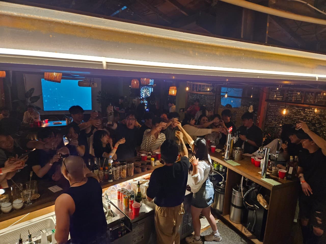 경상북도 경주시 혼술 바(Bar) 진한밤 경주 - 주황색 조명 아래 다찌석에서 사람들이 건배하는 상황이며 1인 카운터에 술병과 붉은색 컵이 배치됨