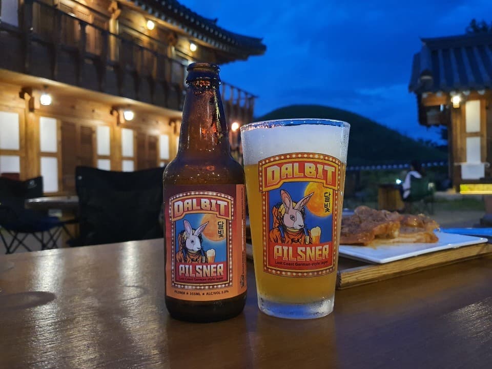 경상북도 경주시 황남거북이 경주 맥주 호프 한옥 야외석 달빛 필스너 병맥주와 거품 가득한 전용잔. 조명 켜진 한옥 배경의 나무 테이블에서 1인 혼술 중인 상황.