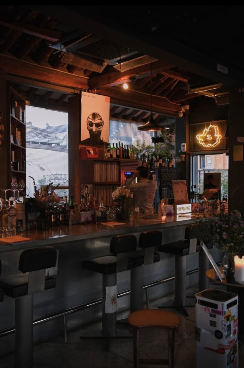 경상북도 경주시 오아시스 경주 황리단길 와인 바의 주황색 조명 아래 나란히 놓인 1인 다찌석. 한옥 서까래와 포스터, 와인병이 진열된 카운터에서 혼술을 즐기는 상황.