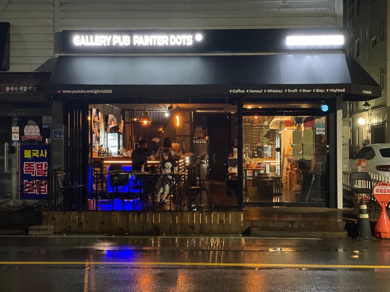 경상북도 경주시 혼술 바 페인터 닷츠 경주. 노란 조명 아래 다찌석과 1인 테이블에 앉아 위스키를 즐기는 상황. 비 내린 밤 거리의 빛이 반사되는 통유리창 너머 매장 내부 전경.