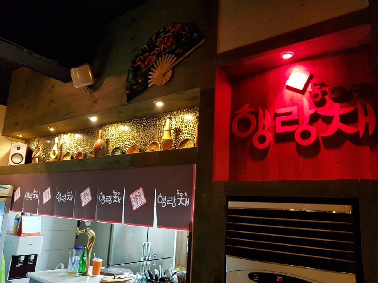 경상북도 포항시 동마실행랑채 포항 맥주 호프. 붉은 조명의 간판 아래 생맥주 탭과 주방 다찌석이 위치한 1인 혼술 공간. 상단에 부채 장식과 도자기가 진열됨.