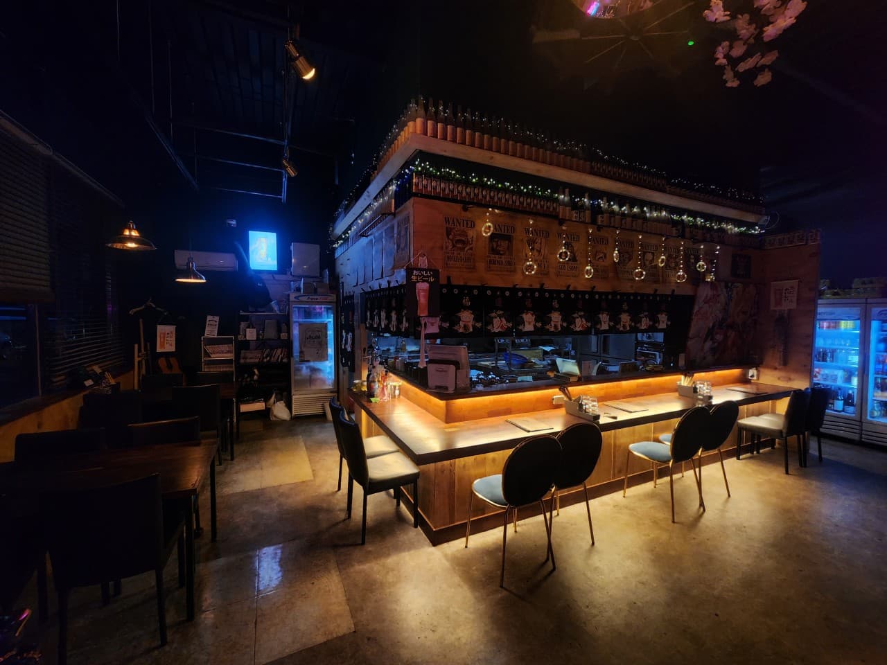 경상북도 포항시 혼술 이자카야 소휴 포항 - 주황색 조명이 비추는 나무 소재 다찌석과 1인 의자가 나란히 배치된 내부. 어두운 배경 속 카운터 테이블과 상단에 진열된 술병.