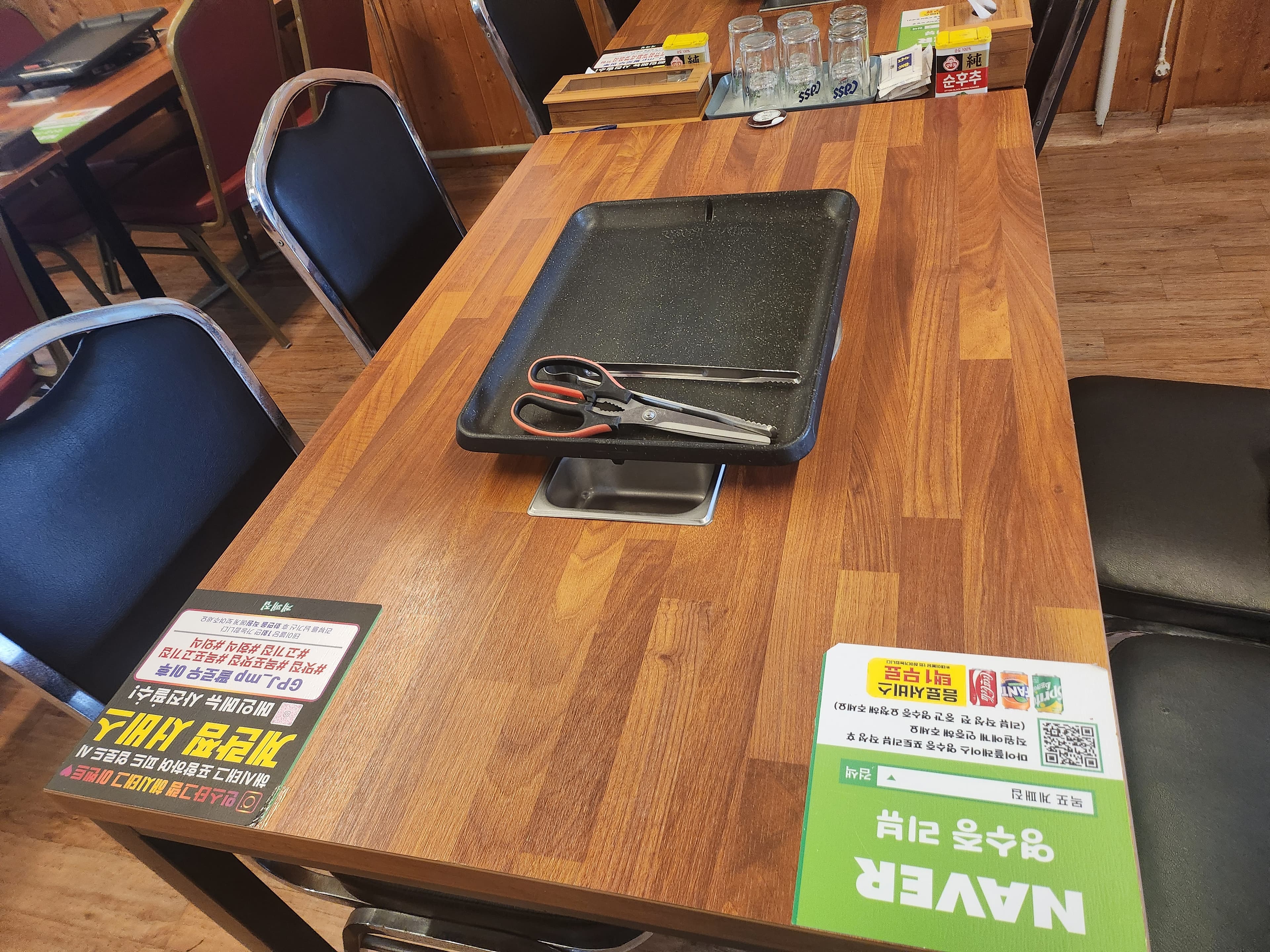 전라남도 목포시 목포 계패집의 나무 테이블 위 검정 사각 불판과 금속 집게, 가위. 혼밥을 위한 1인석에 맥주잔과 리뷰 이벤트 안내문이 놓인 기타 매장 세팅 상황