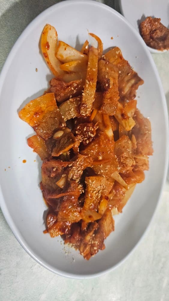 부산광역시 동구 솔잎국수 초량 포장마차 다찌석에서 즐기는 1인 혼술. 하얀 접시에 담긴 매콤한 고기 볶음과 깨의 질감이 강조된 안주 차림.
