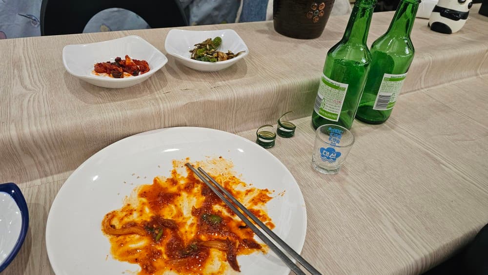 부산광역시 동구 신촌포차 초량 포장마차 - 조명 아래 나무 다찌석에 놓인 빨간 양념의 1인 안주 접시와 빈 소주병. 카운터석에서 혼술을 마친 상황.