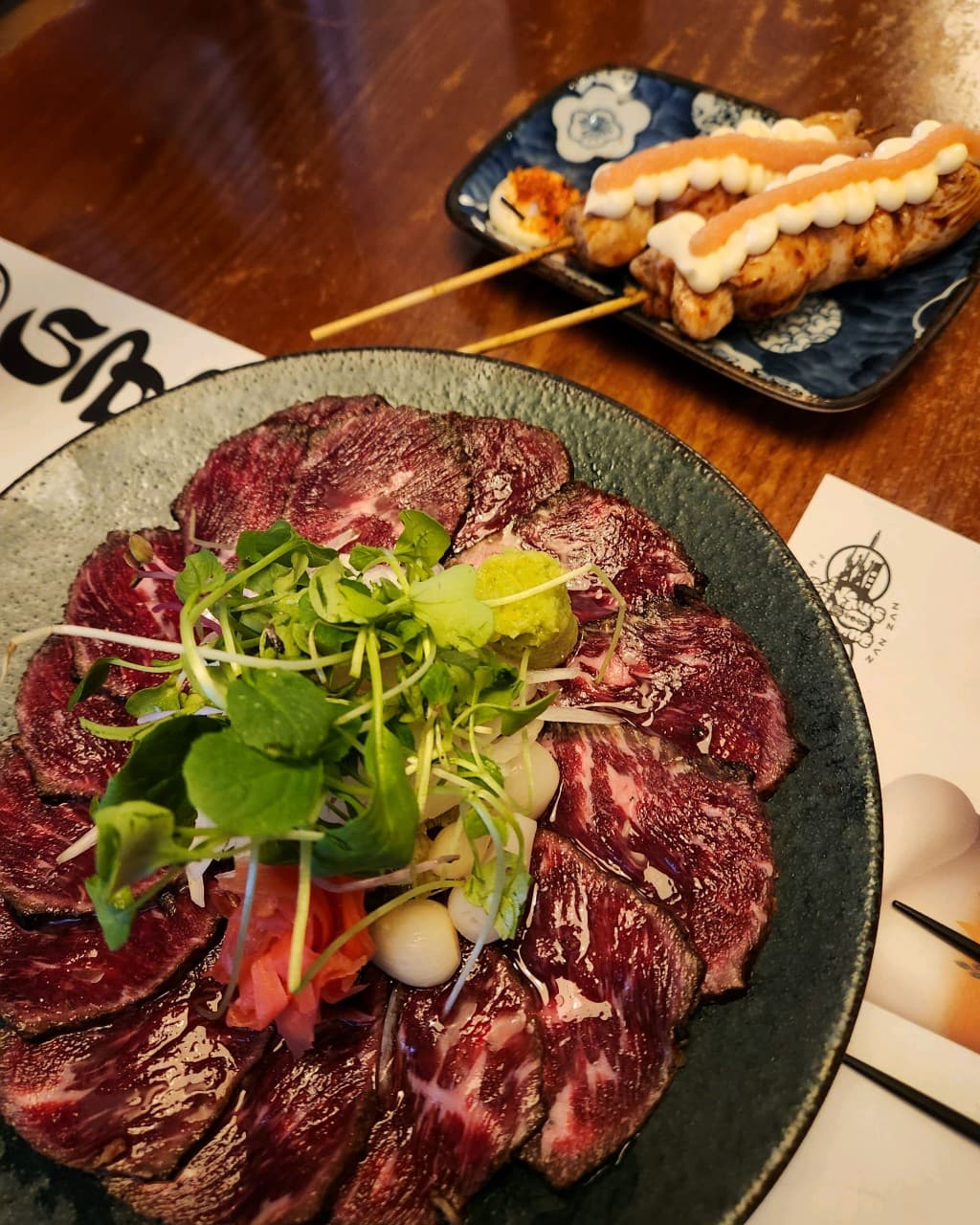 경기도 안양시 잔잔 범계역점 이자카야 나무 다찌석 위 소고기 타타키와 명란 꼬치. 노란 조명 아래 혼술 상황을 보여주는 1인 메뉴와 선명한 육질의 상차림.