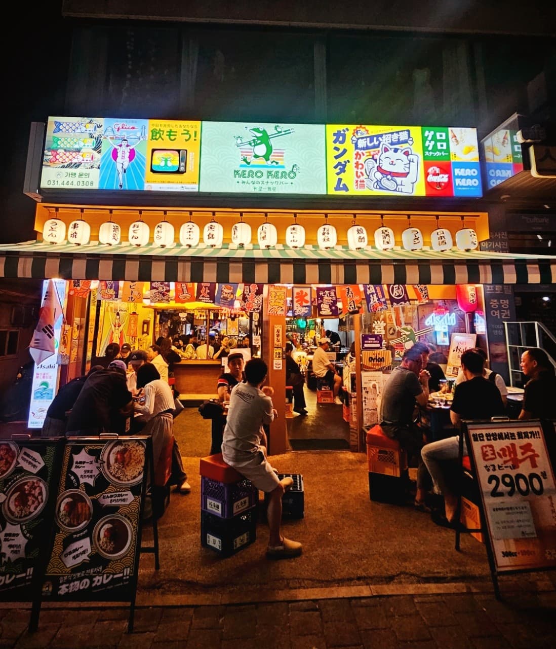 경기도 안양시 이자카야 케로케로 안양점 경기 - 노란 제등 조명 아래 야외 플라스틱 좌석과 내부 다찌석에서 1인 혼술 중인 사람들과 일본풍 간판 묘사