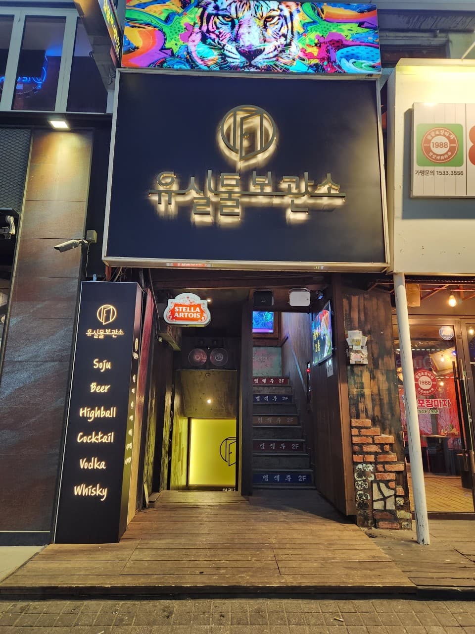 경기도 안양시 유실물보관소 경기 안양 입구. 혼술 메뉴인 하이볼과 위스키가 적힌 간판 옆으로 나무 데크와 조명이 켜진 계단, 상단에 호랑이 일러스트가 있는 건물 외관.