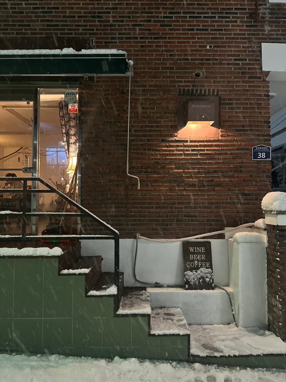 경기도 성남시 아트살롱404 맥주 호프 - 눈 내리는 붉은 벽돌 건물과 주황색 조명 아래 창가 혼술 좌석. 입구 계단과 와인 맥주 표지판이 배치됨.