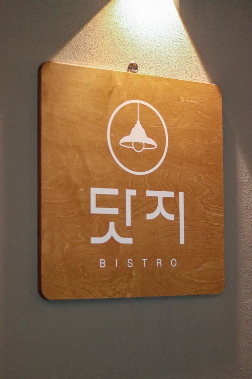 경기도 구리시 혼술 요리주점 닷지 - 핀 조명이 수직으로 비추는 나무 간판과 다찌석 비스트로 로고 및 벽면의 질감이 드러난 입구 공간