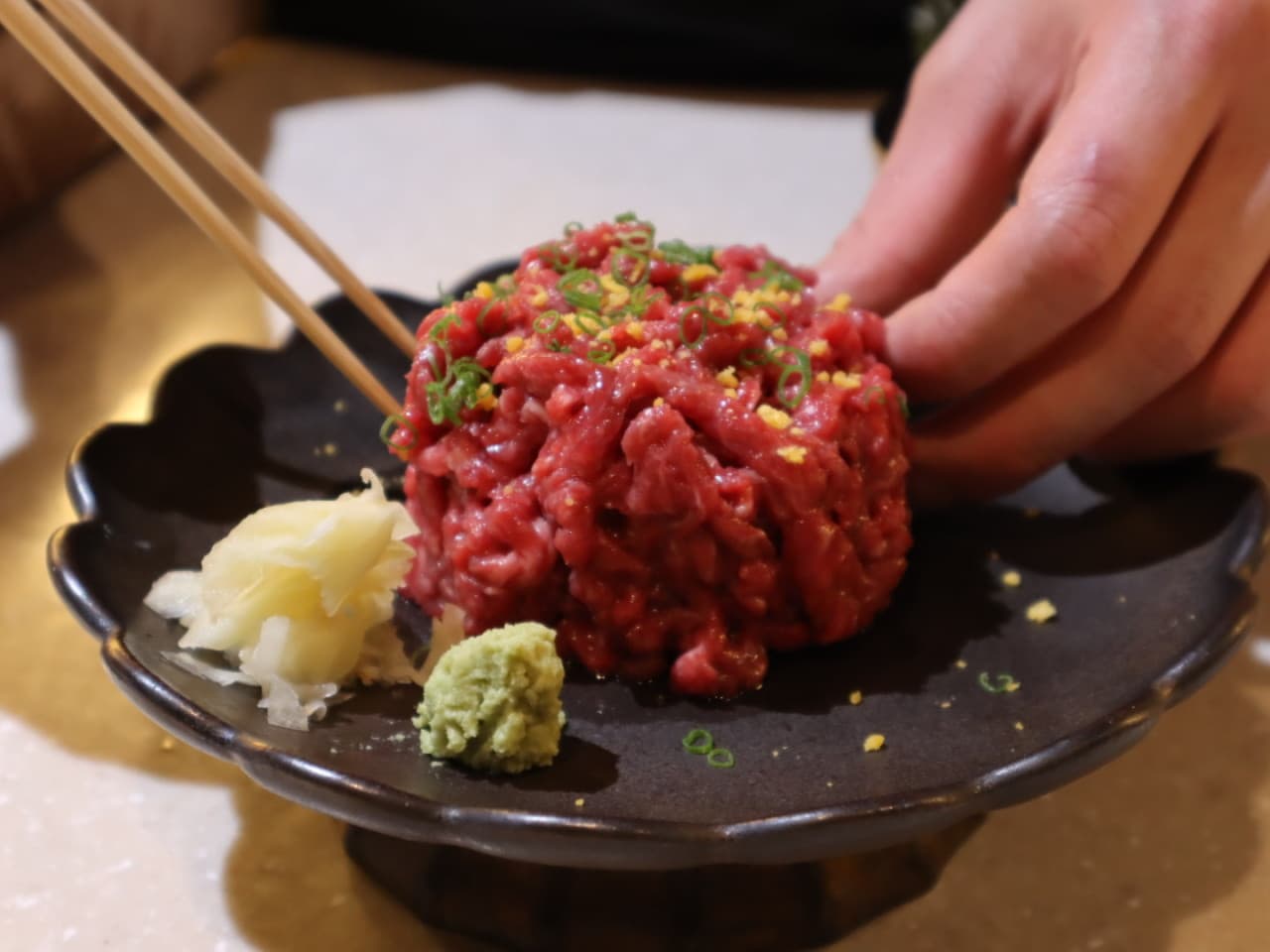 서울특별시 광진구 혼술 이자카야 류지로 다찌석. 따뜻한 조명 아래 검은 접시에 담긴 선홍빛 육회와 와사비를 젓가락으로 집는 1인 식사 상황.
