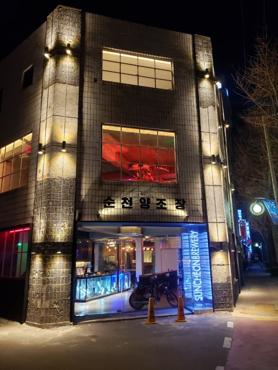 전남광주통합특별시 순천시 순천양조장 맥주 호프 - 조명 아래 다찌석과 1인 테이블이 있는 건물 외관. 푸른 네온사인과 내부 바이크가 혼술을 위한 공간 구성을 보여줌.