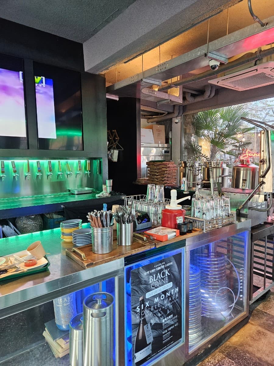 전남광주통합특별시 순천시 순천양조장 맥주, 호프 - 초록색 네온 조명 아래 스테인리스 다찌석과 정돈된 식기류, 브루어리 설비가 배치된 1인 혼술 공간