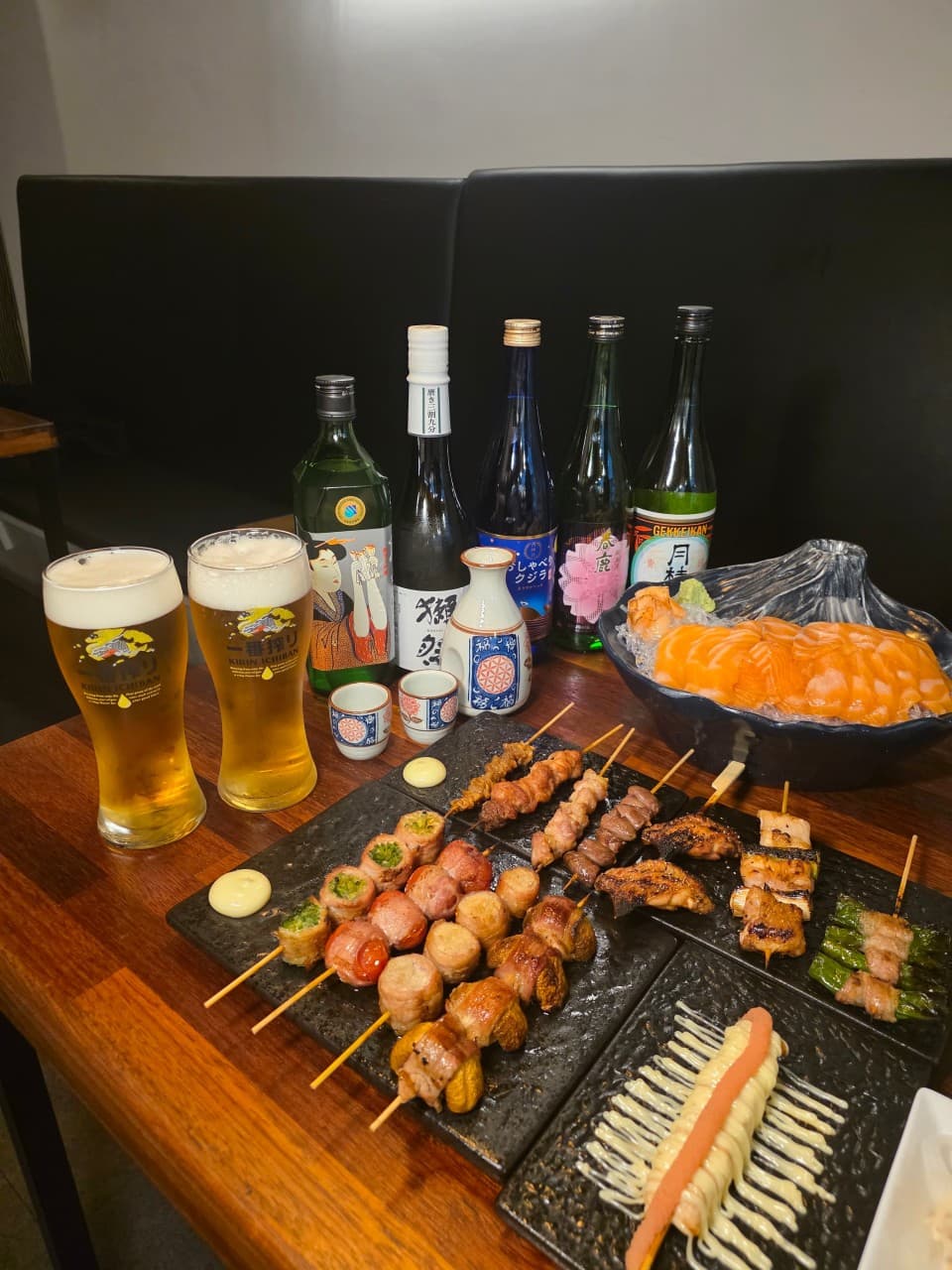 전남광주통합특별시 순천시 야키토리 신지야 순천 이자카야 혼술. 나무 테이블 위 노릇한 닭꼬치와 연어 사시미, 생맥주와 사케 병이 나열된 조명 아래 1인 식사 상황.