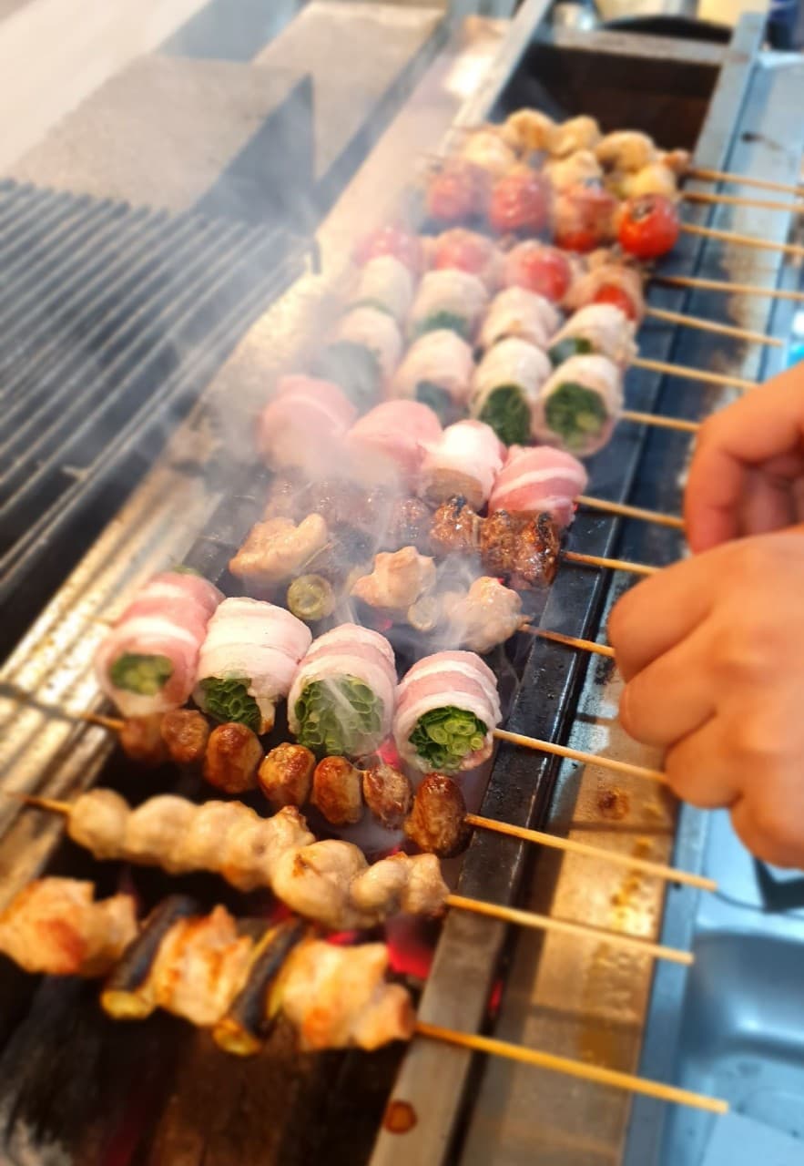 전남광주통합특별시 순천시 야키토리 신지야 순천 이자카야 다찌석 혼술. 석쇠 위 연기와 함께 익어가는 닭꼬치와 베이컨 채소말이의 질감이 보이는 상황.