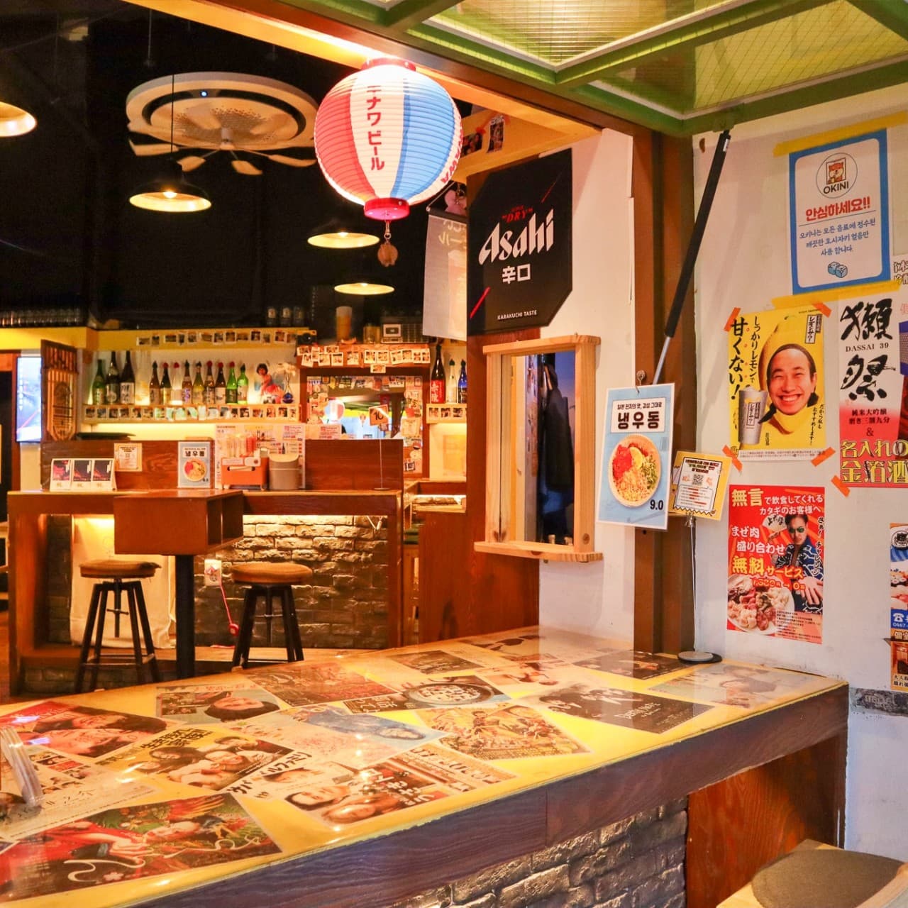 전남광주통합특별시 목포시 이자카야 오키니 전남의 붉은 제등 아래 일본풍 포스터가 붙은 다찌석. 1인 혼술 바 좌석과 노란 조명이 비치는 매장 내부 전경.