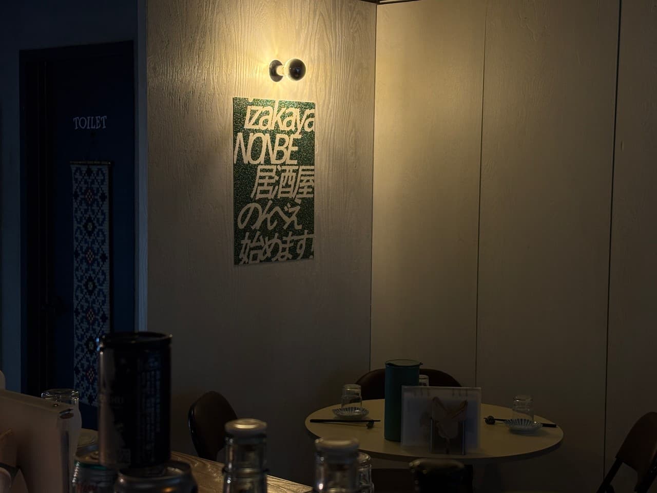 서울특별시 마포구 논베 서울 이자카야. 벽면 포스터 아래 노란 조명과 다찌석, 1인 방문이 가능한 원형 테이블이 있는 내부. 혼술에 적합한 조용하고 정갈한 식기 세팅이 보임.