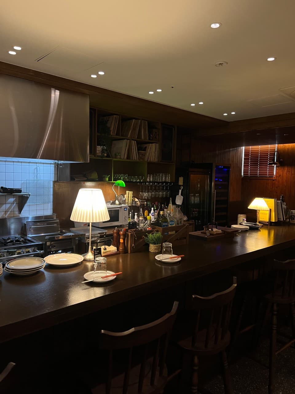 서울특별시 강서구 혼술 이자카야 바드 서울 - 낮은 조도 아래 긴 원목 다찌석과 1인 식기 세팅. 스탠드 조명이 비추는 카운터석과 배경의 레코드 선반이 위치함.