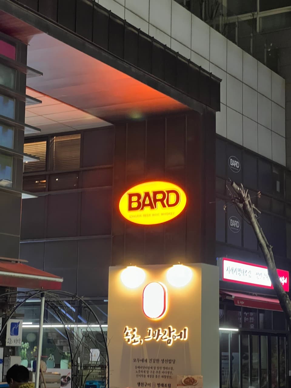 서울특별시 강서구 이자카야 바드 서울의 노란색 원형 간판과 야간 조명. 다찌석이 마련된 1인 혼술 장소의 외경을 보여주는 입구와 건물 외벽 요소.