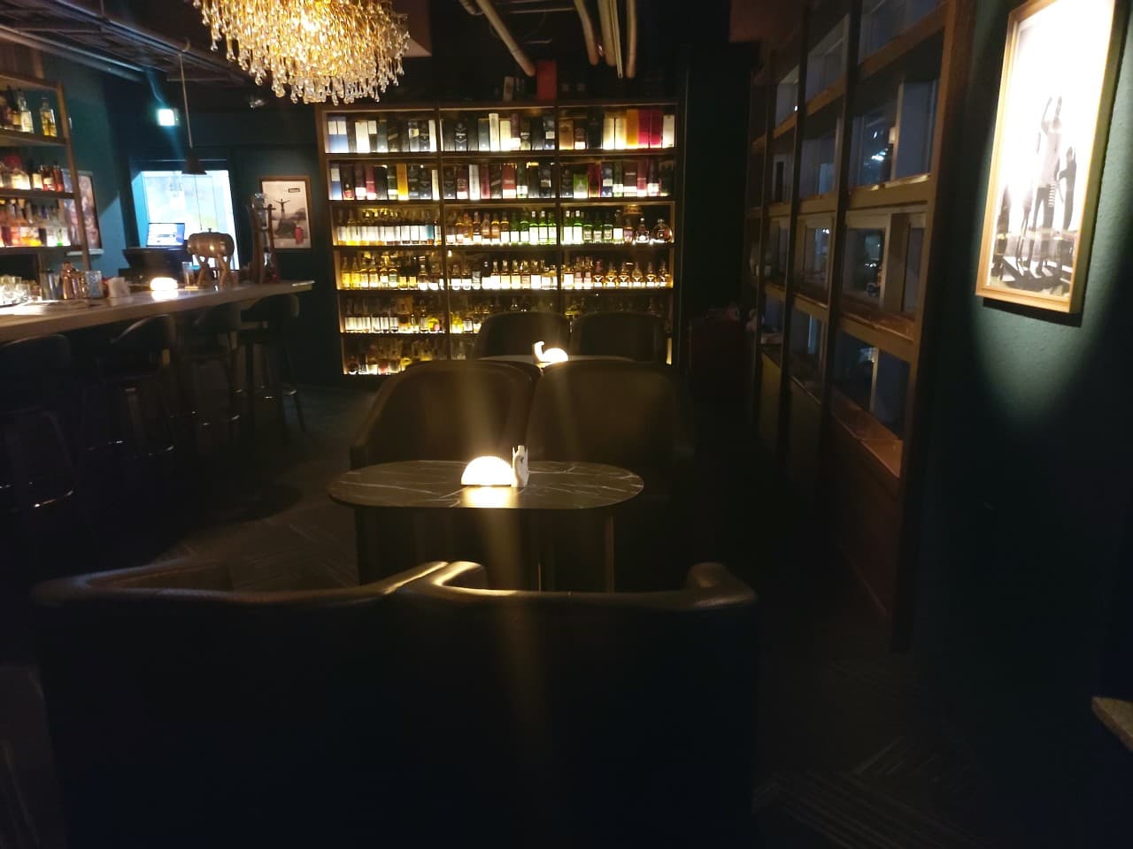 서울특별시 강서구 깔루아 서울 바(Bar)의 샹들리에 조명 아래 배치된 다찌석과 1인 좌석. 벽면 진열장을 가득 채운 위스키 병의 질감과 테이블 위 개별 램프가 혼술 상황을 연출함.