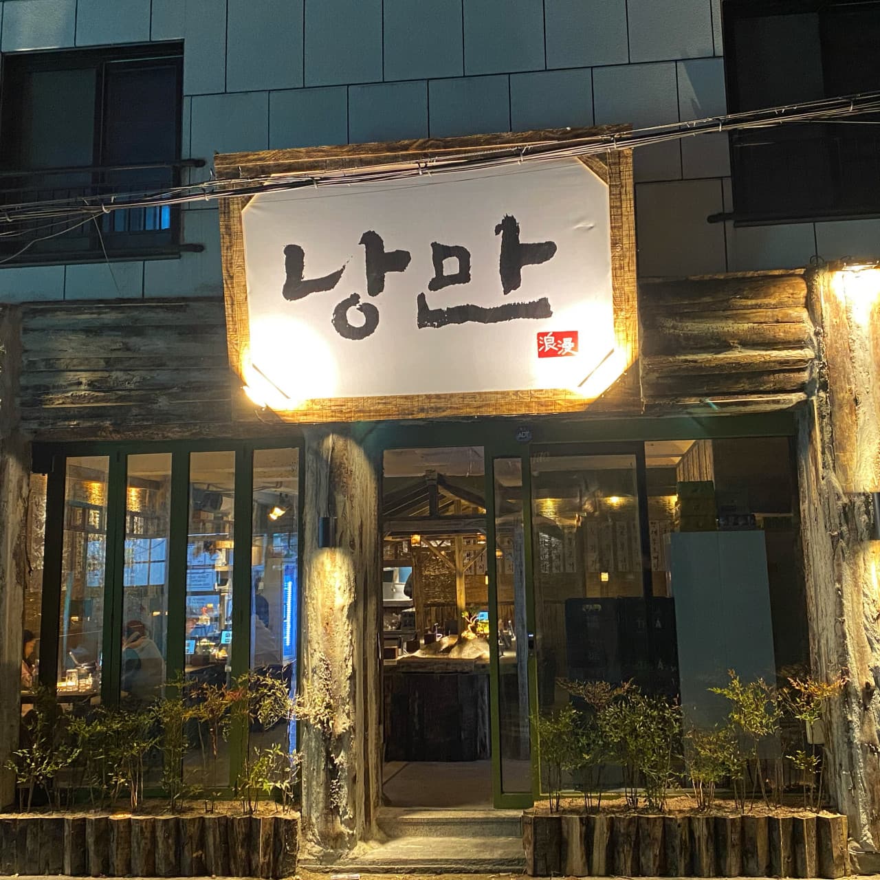 대전광역시 서구 요리주점 낭만짚불구이 대전괴정점. 나무 질감 외벽과 노란 조명이 켜진 입구, 유리창 너머로 1인 다찌석과 목조 구조물이 보이는 혼술 하기 적합한 분위기의 매장.