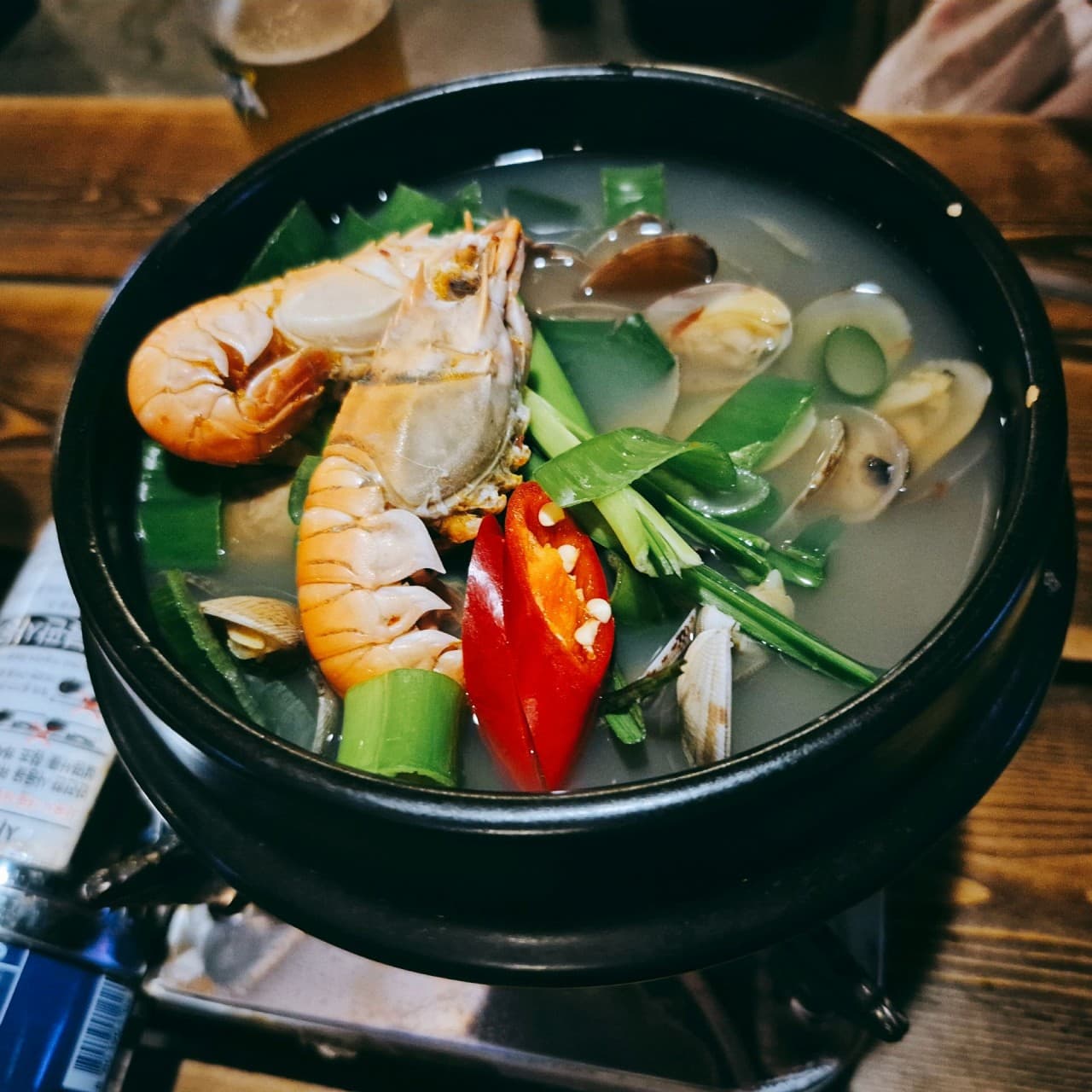 대전광역시 서구 요리주점 낭만짚불구이 대전괴정점. 은은한 조명 아래 나무 테이블 다찌석에서 혼술로 즐기는 딱새우 조개탕. 뚝배기에 담긴 통통한 새우와 대파, 조개의 질감이 강조됨.