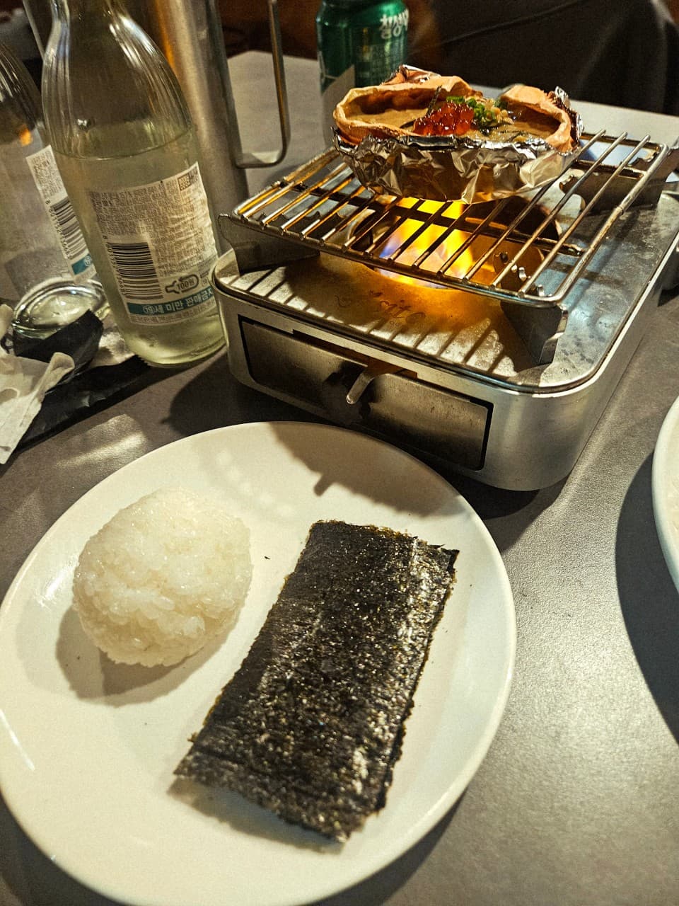 대전광역시 유성구 이자카야 바시트 대전 1인 다찌석. 화로 불꽃 위 게딱지 요리와 연어알, 김과 밥이 차려진 혼술 상황. 투명한 술병과 캔음료가 놓인 식탁.