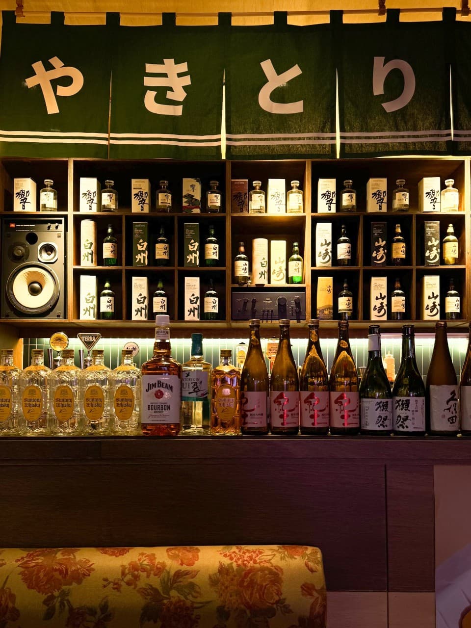 대전광역시 서구 이자카야 야키토리동작 대전가수원점. 노란 조명 아래 위스키와 사케 병이 정렬된 선반 앞 나무 다찌석에서 즐기는 1인 혼술 상황.