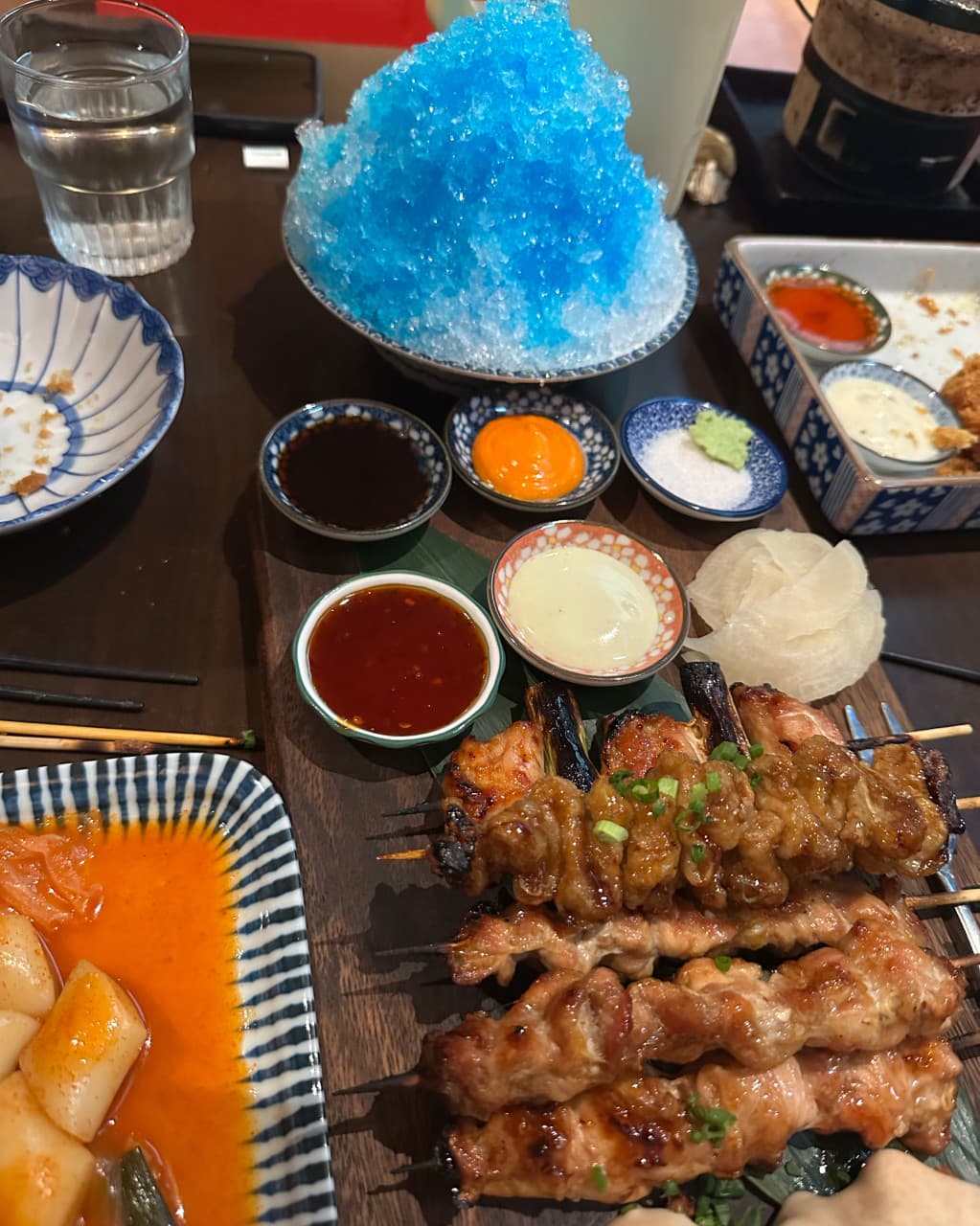대전광역시 서구 야키토리동작 대전가수원점 이자카야 다찌석 조명 아래 차려진 1인 상차림. 윤기 나는 닭꼬치와 파란색 빙수, 떡볶이 질감이 돋보이는 혼술 상황.