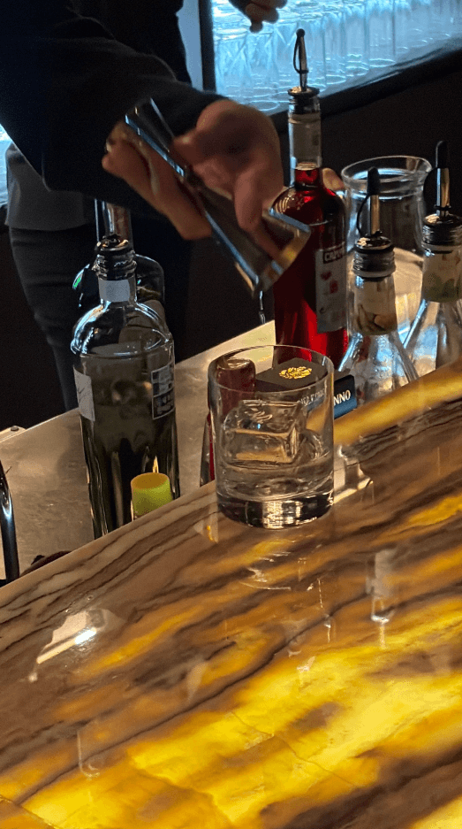 대전광역시 유성구 루미테라 대전 바(Bar) - 노란 조명의 다찌석 위 투명한 각얼음 잔과 술을 조제하는 바텐더가 있는 1인 혼술 공간