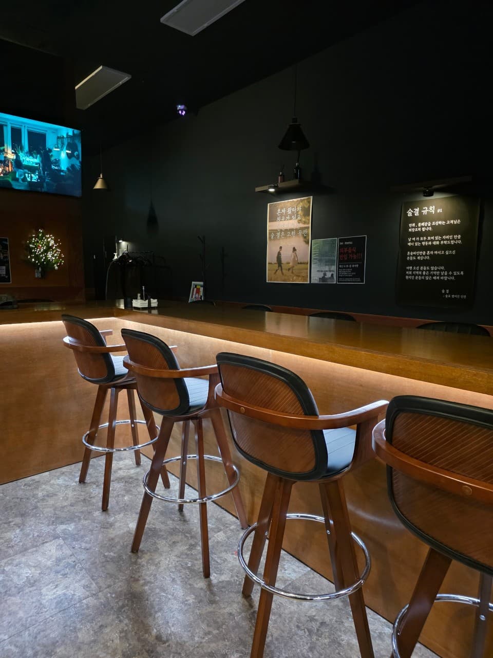 전남광주통합특별시 목포시 술결 목포혼술바 전남 바(Bar)의 나무 소재 긴 다찌석과 1인용 의자. 어두운 벽면과 대비되는 하단 간접 조명이 비치는 공간에서 혼술을 즐기는 상황을 묘사함.
