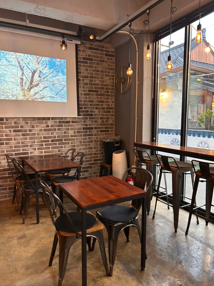경상북도 경주시 황남맥주 경북 맥주 호프. 벽돌 벽과 프로젝터가 설치된 실내, 창가 1인 바 좌석과 전구 조명이 비추는 창 밖 한옥 배경의 혼술 상황.