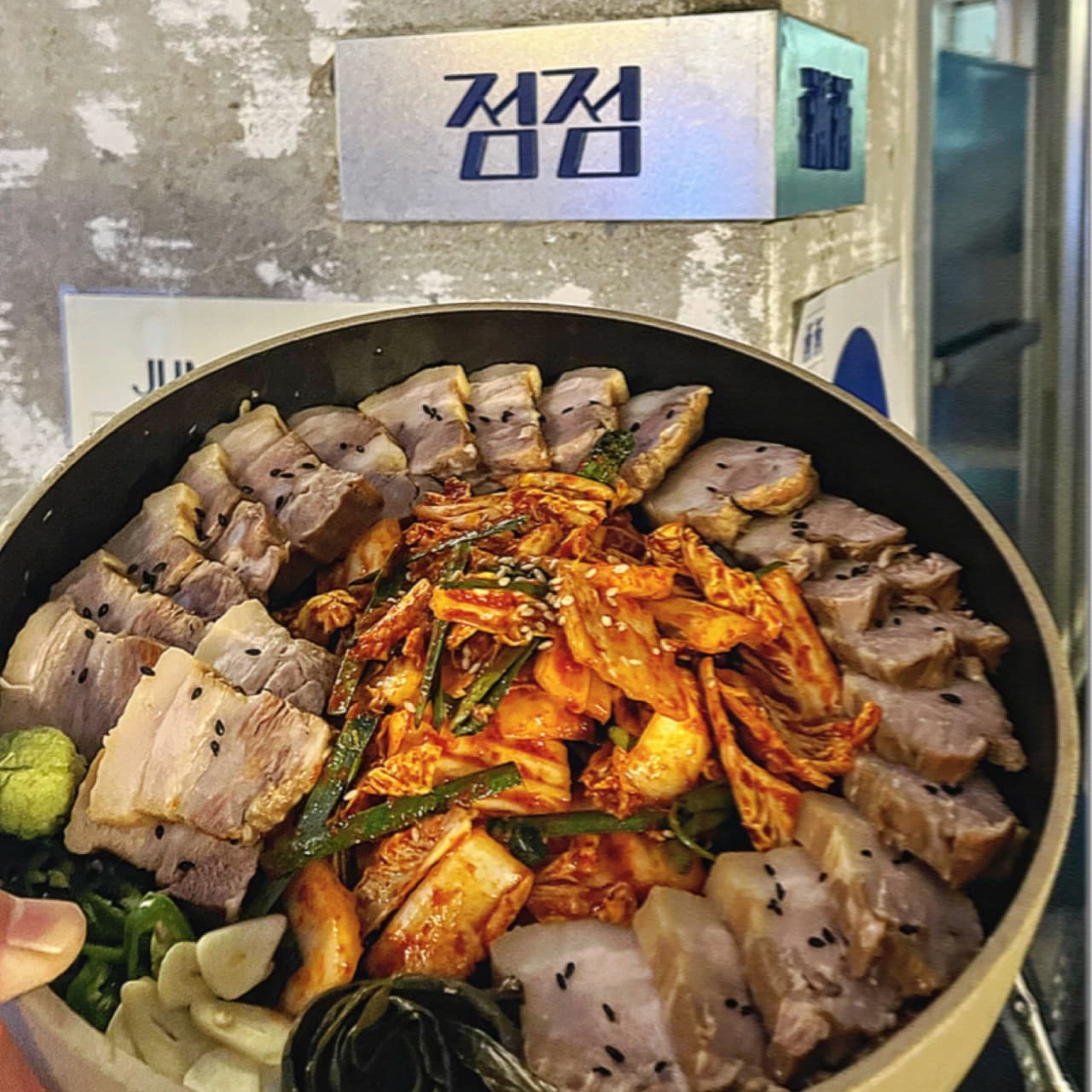 경상북도 경주시 요리주점 점점 경북 다찌석에 앉아 혼술로 즐기는 1인 수육 보쌈. 조명 아래 검은 깨가 뿌려진 고기 질감과 붉은 김치가 노출 콘크리트 배경과 어우러짐.